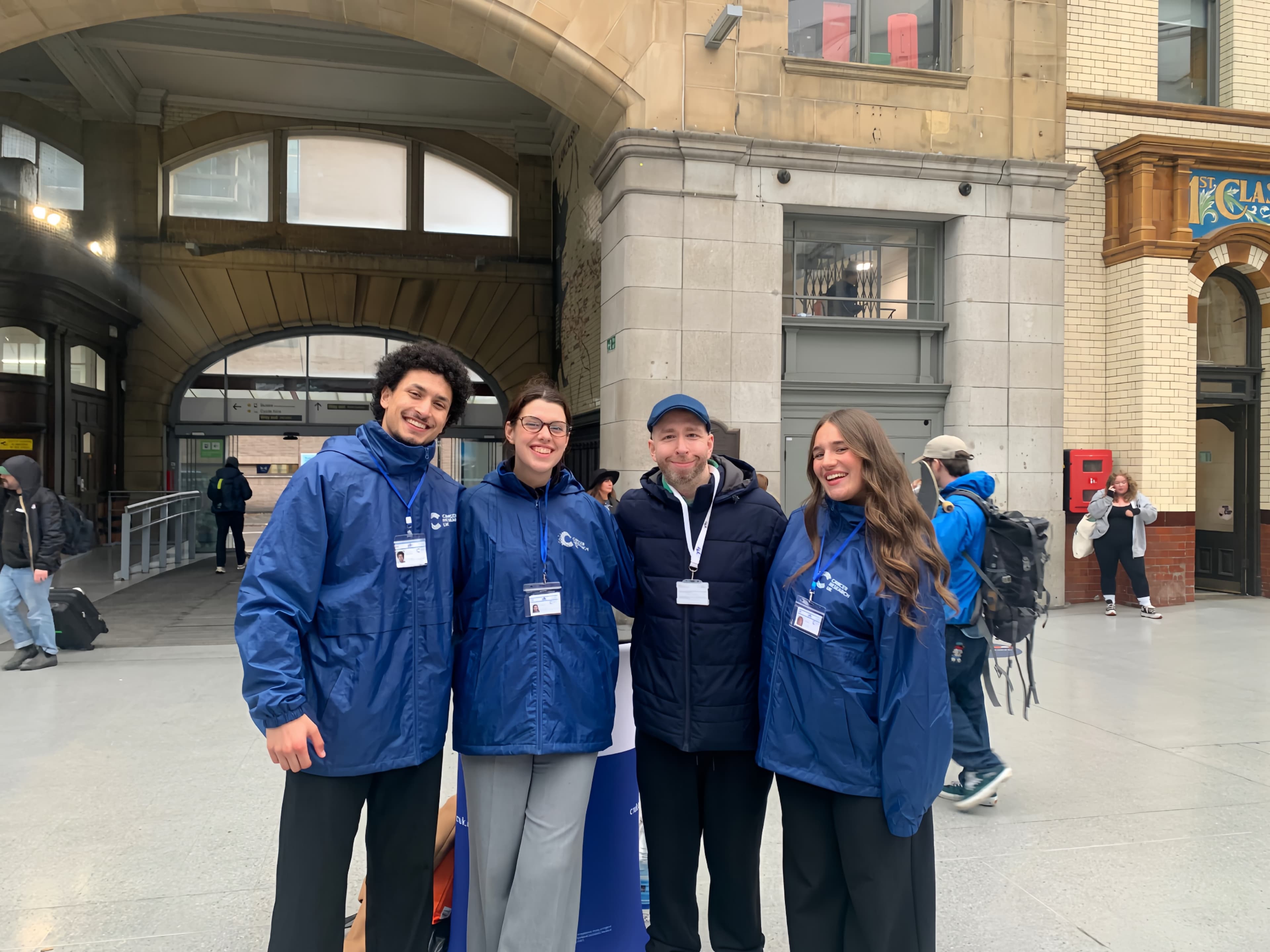 Four smiling people in blue jackets and lanyards stand together in a large arched building.