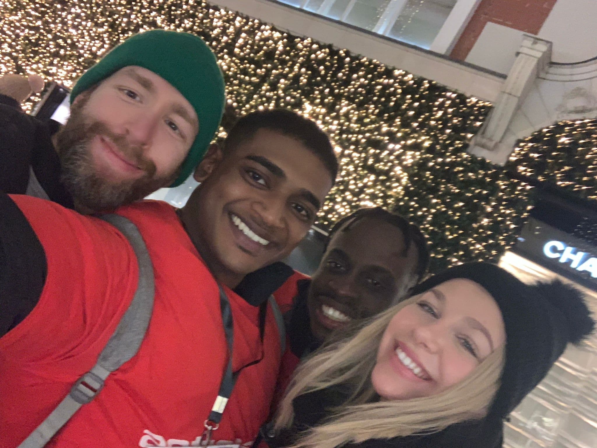 Four diverse friends smile for a selfie against a backdrop of glowing festive fairy lights.