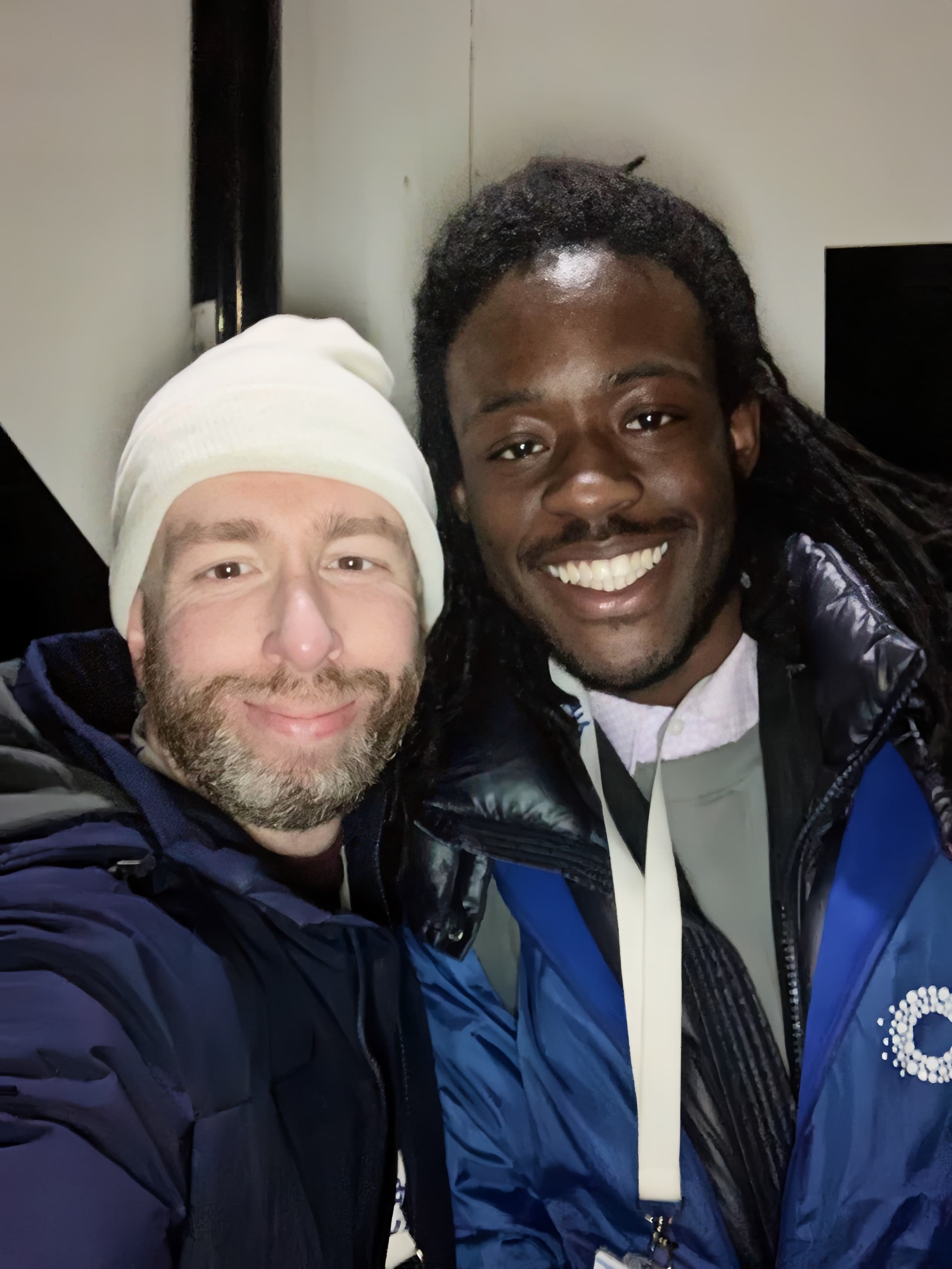 Bearded man in white beanie and smiling man with dreadlocks pose for a selfie together.