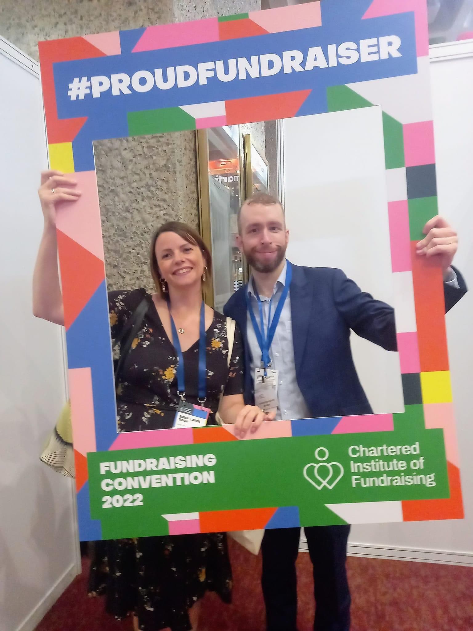 Two smiling people pose behind a colorful #PROUDFUNDRAISER frame at the Fundraising Convention 2022.