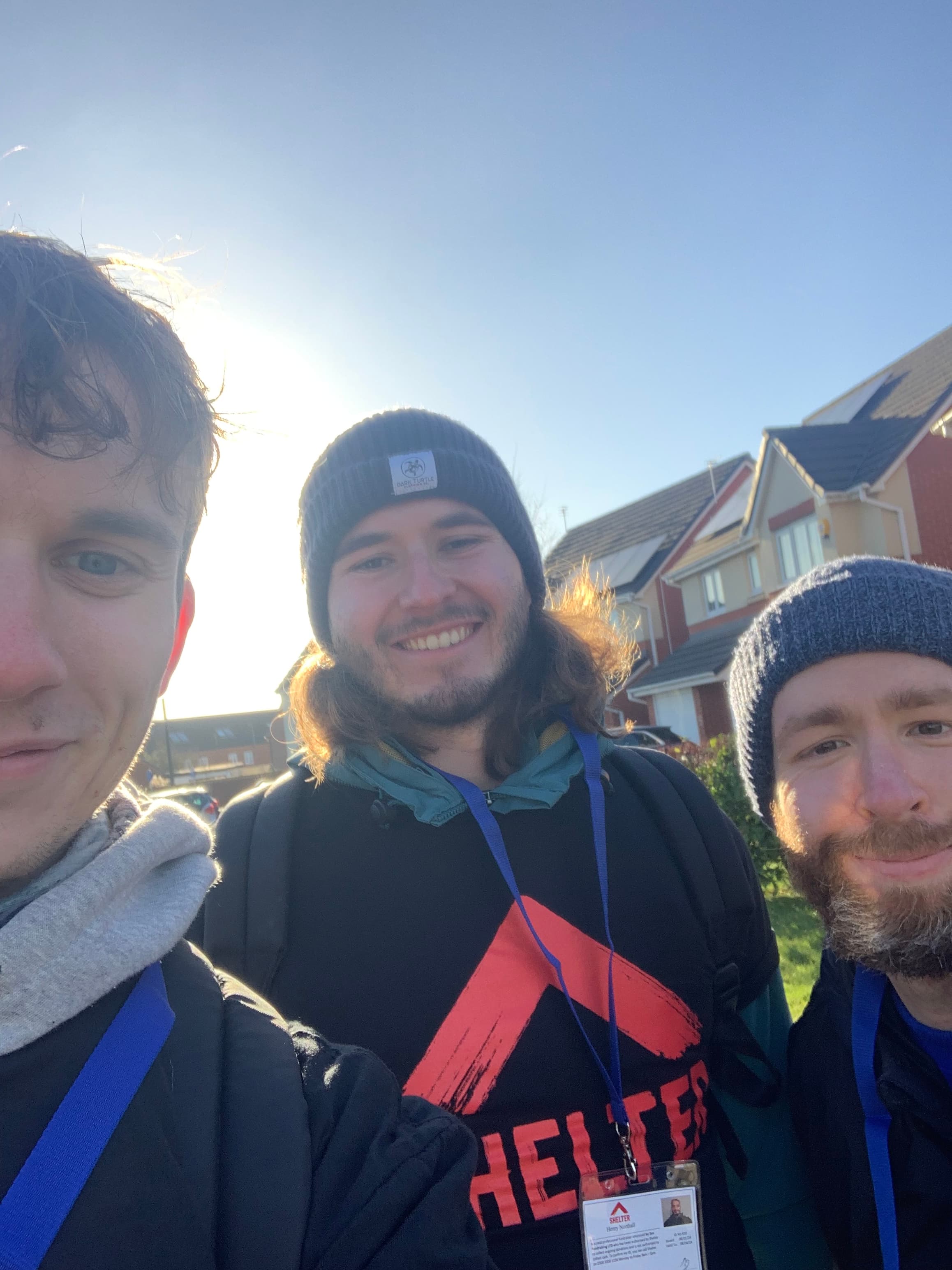 Three men smiling for a sunny outdoor selfie, wearing beanies and lanyards in a neighborhood.