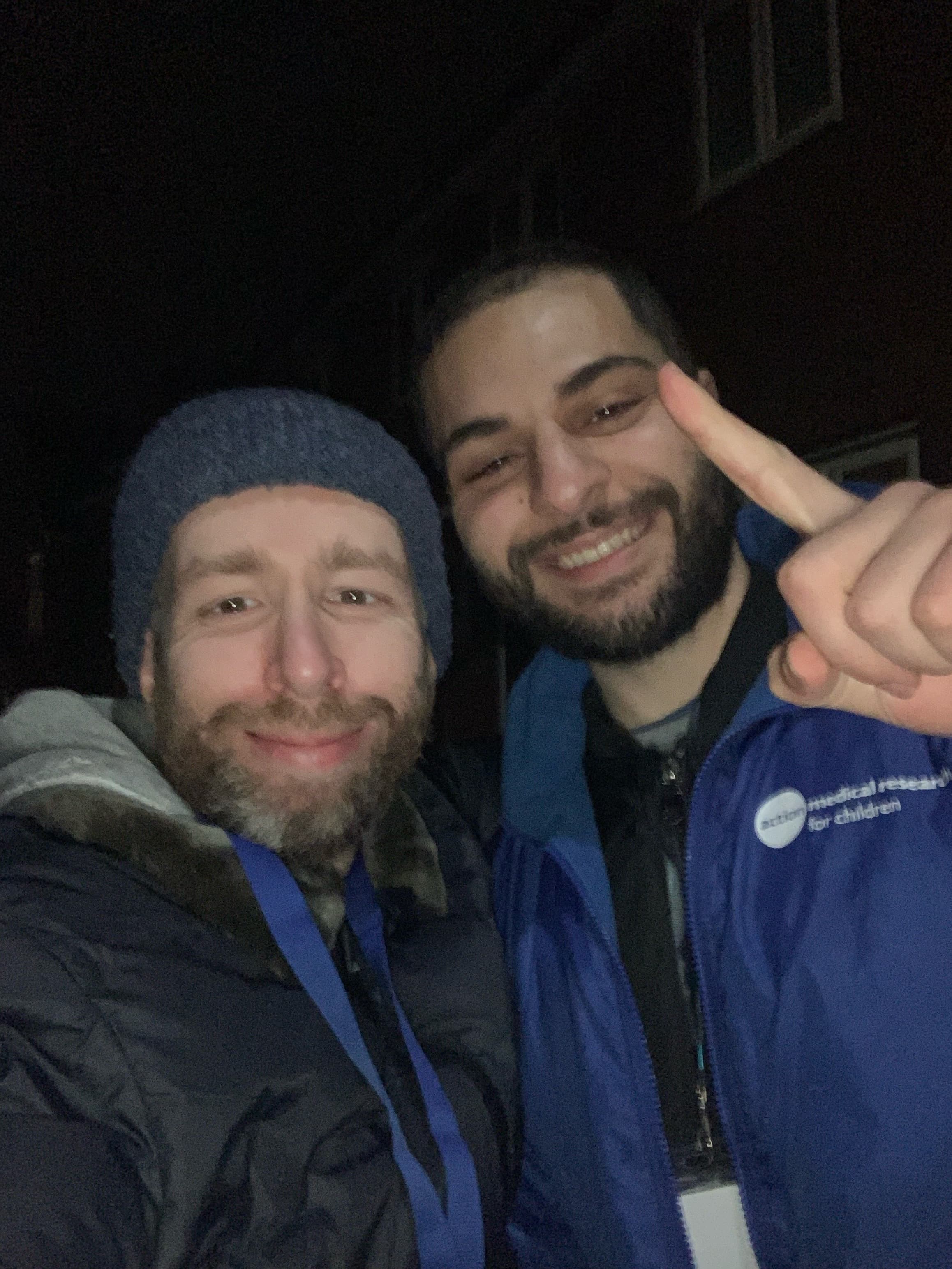 Two men smiling for a night selfie, one wearing a beanie and another pointing up.