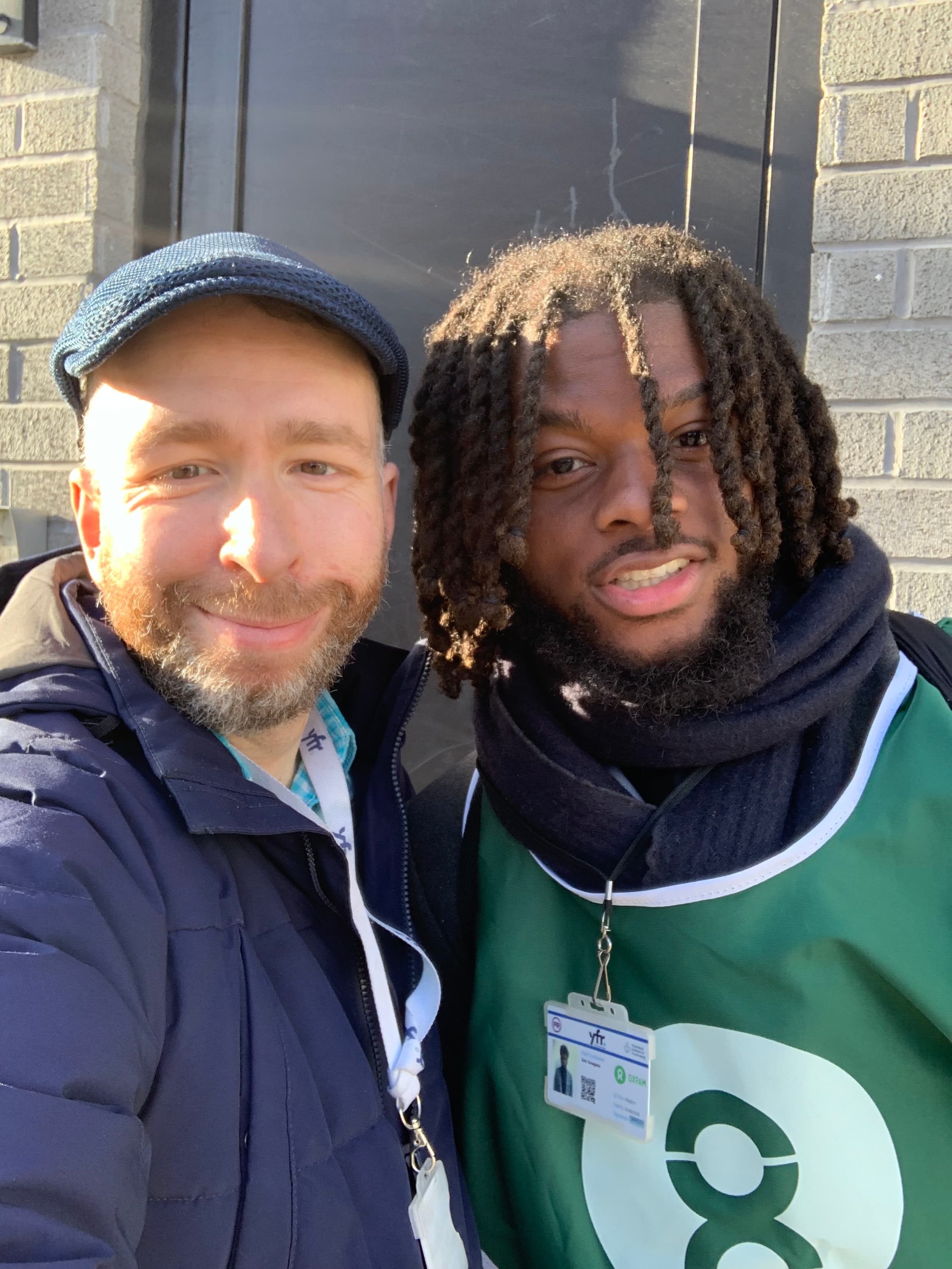 Two smiling men, one with dreadlocks in an Oxfam vest, take a selfie outdoors.