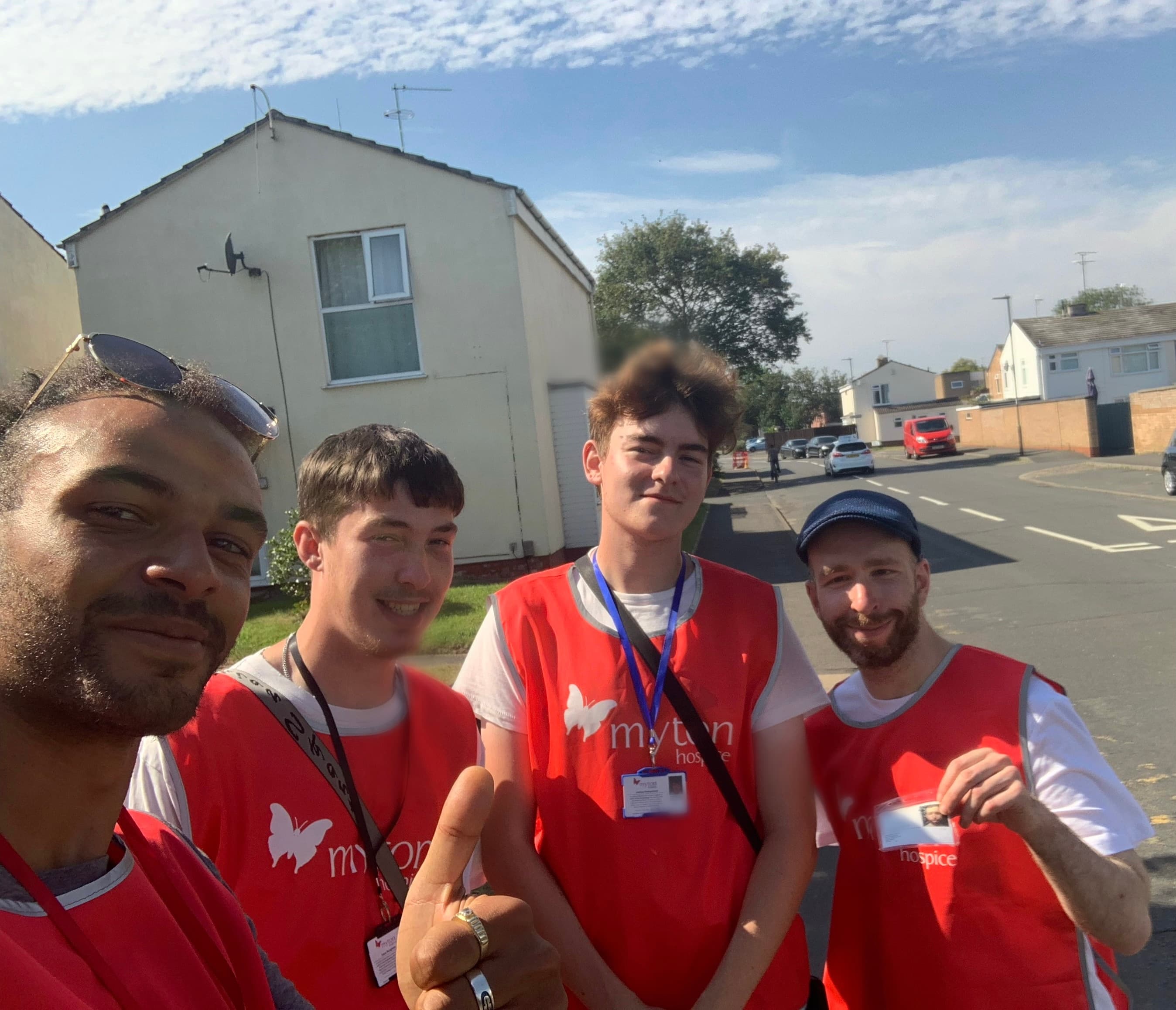Four men in orange Myton Hospice vests smile for a selfie on a sunny street.
