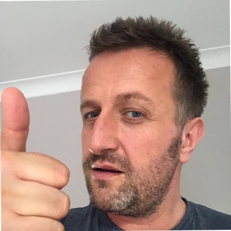 Man with spiky hair and a beard giving a thumbs-up sign in a selfie.